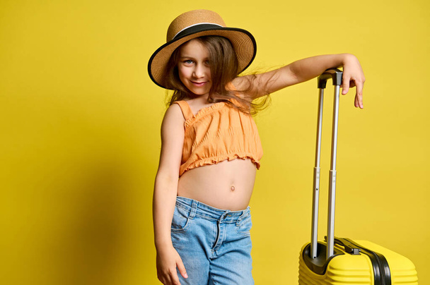 Boldog gyermek lány aranyos mosolyog a kamera előtt, pózol elegáns sárga bőrönd, öltözött fényes narancssárga felső, kék farmer és szalma kalap, elszigetelt sárga háttér. Utazás, gyerekek és nyári koncepció - Fotó, kép