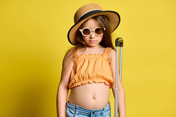 Caucasian adorable child girl, little tourist passenger in stylish summer wear, straw hat and sunglasses, posing with suitcase, looking confidently at camera, isolated on yellow background. Tourism - Photo, Image