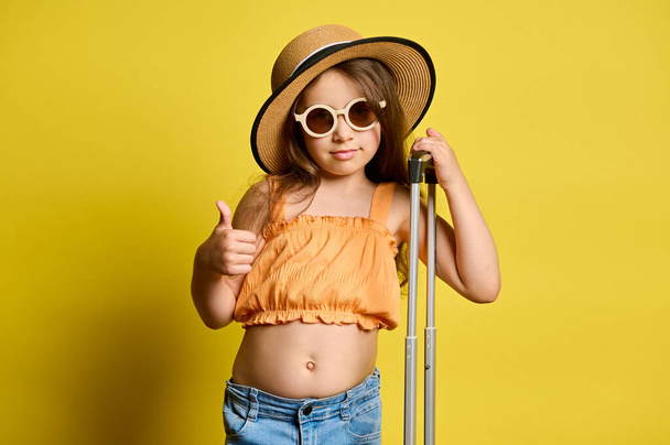 Happy little child girl, holiday maker, in stylish sunglasses, summer straw hat and orange top, gestures with thumb up, looking at camera, isolated over yellow color background. Trip. Travel. Journey - Foto, imagen