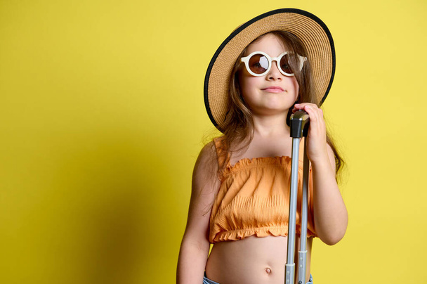 Valkoihoinen suloinen tyttö aurinkolaseissa, olkihattu ja oranssi toppi, poseeraa käsimatkatavaroiden kanssa, eristettynä keltaisen studiotaustan päällä. Kopioi mainostila. Matkailu. Matkustaa. Tripin konsepti. Lentomatka - Valokuva, kuva