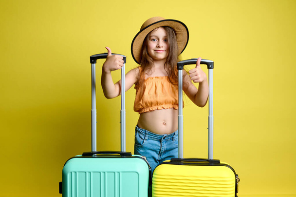 Mosolygó kis turista utas elegáns alkalmi nyári ruhák, gesztusok hüvelykujjal felfelé, álló sárga és világos zöld polikarbonát, műanyag bőröndök, elszigetelt stúdió háttér - Fotó, kép