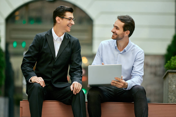 Cheerful discussion. Two businessmen, coworkers sitting on bench outside the office, looking on laptop and working. Concept of business, career development, ambitions, success, office lifestyle - Photo, image