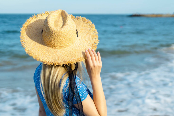 Mujer en vestido con sombrero de paja caminando por el mar en el día soleado, vista trasera.  - Foto, Imagen