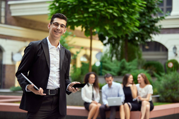 Focus on smiling, confident businessman standing with laptop and mobile phone outside office. Blurred people on background. Business, career development, ambitions, success, office lifestyle concept - 写真・画像