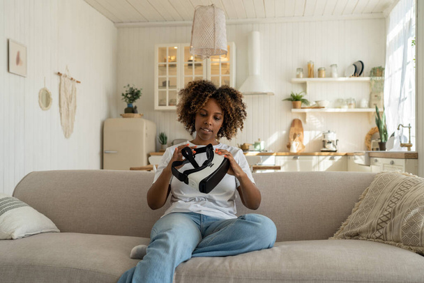 Захоплена афро-американська жінка, дивлячись на свій перший VR навушники і відчуваючи себе в захваті, щоб зануритися, сидячи на дивані в вітальні. Цікава чорна леді геймер тримає футуристичні окуляри віртуальної реальності - Фото, зображення