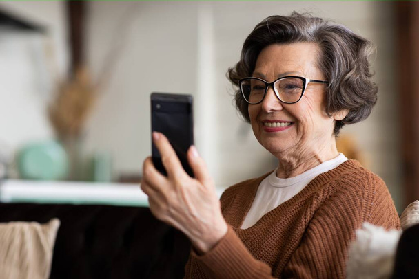 Gelukkig volwassen vrouw van middelbare leeftijd genieten van het gebruik van mobiele apps, sms 'en, zitten op de bank, glimlachen senior vrouw met smartphone, kijken naar mobiele telefoon scherm, browsen social media thuis. Hoog - Foto, afbeelding