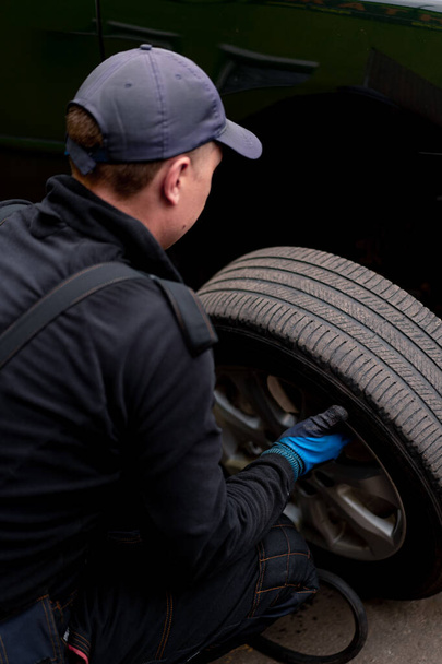 Автомеханик в кепке меняет колесо с черного автомобиля с помощью дрели в шинном цехе на улице, подробно описывая ремонт автомобиля - Фото, изображение