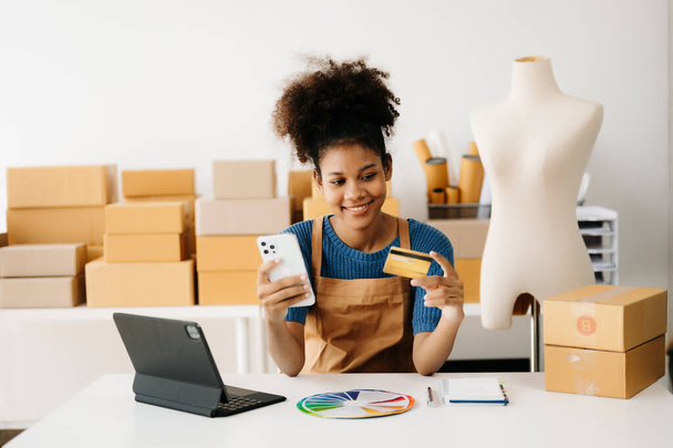 Entrepreneur owner. African American woman using smartphone and tablet at desk, taking receive and checking online purchase shopping order to preparing pack product boxes  - Photo, image
