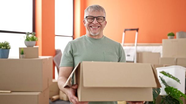Grauhaariger Mann mittleren Alters lächelt zuversichtlich und hält Paket im neuen Zuhause - Foto, Bild