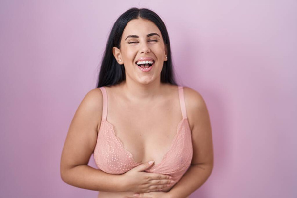 Молода іспаномовна жінка в рожевому бюстгальтері посміхається і сміється голосно, тому що смішний божевільний жарт з руками на тілі.  - Фото, зображення