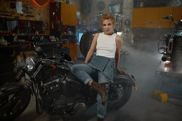 ガレージのオートバイに座って魅力的な女性のバイカー。ワークショップでカメラのためにポーズをとるかなり女性機械工 - 写真・画像