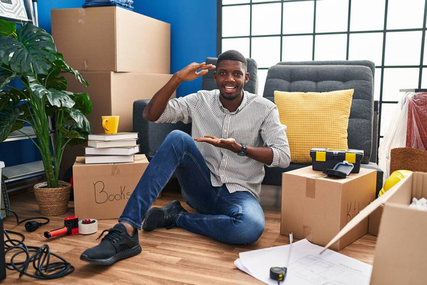Afrikkalainen amerikkalainen mies istuu lattialla uudessa kodissa eleiden kädet osoittavat iso ja suuri koko merkki, mitta symboli. hymyillen katsoen kameraan. mittauskäsite.  - Valokuva, kuva