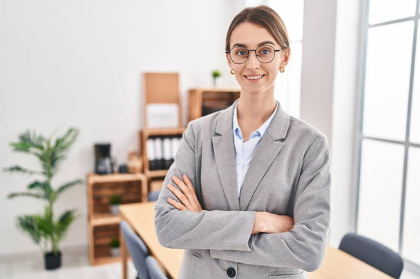 Молода біла жінка бізнесмен посміхається впевнено стоячи з обіймами схрестив жест в офісі
 - Фото, зображення