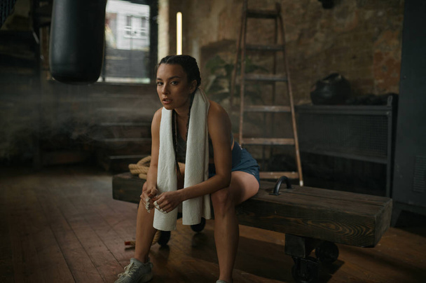 Χαλαρωμένη γυναίκα πυγμάχος που αναπαύεται στον πάγκο με πετσέτα στο λαιμό. Νεαρό κουρασμένο κορίτσι σε αθλητικά είδη κάθεται πάνω από το παλιό γυμναστήριο grungy εσωτερικό - Φωτογραφία, εικόνα