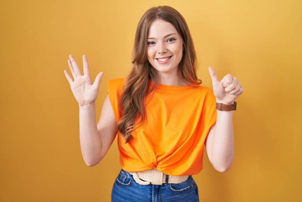 黄色い背景の上に立っている白人女性は自信と幸せに微笑んでいる間指の番号6を指で指で指を指さす..  - 写真・画像