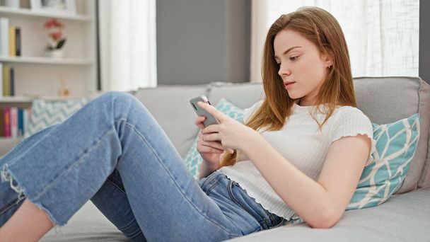 Fiatal szőke nő használ smartphone fekvő kanapén otthon - Fotó, kép