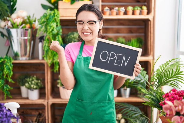 Hispanic young woman working at florist holding open sign screaming proud, celebrating victory and success very excited with raised arm  - Photo, Image