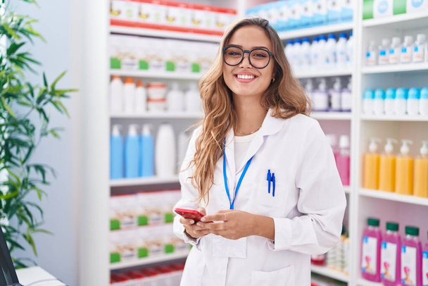 Young beautiful hispanic woman pharmacist using smartphone working at pharmacy - 写真・画像