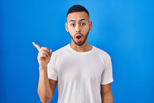 Young hispanic man standing over blue background surprised pointing with finger to the side, open mouth amazed expression.  - Foto, imagen