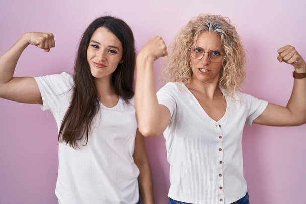 Мать и дочь стоят вместе на розовом фоне, показывая мускулы рук, гордо улыбающиеся. фитнес концепция.  - Фото, изображение