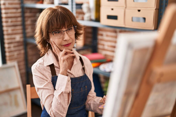 Μέση ηλικία γυναίκα καλλιτέχνης αναζητούν κλήρωση με την έκφραση αμφιβολία στο στούντιο τέχνης - Φωτογραφία, εικόνα
