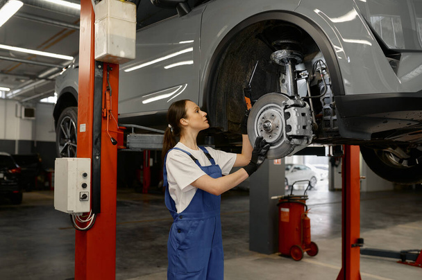 Woman auto mechanic fixing wheel hub or disc brake of raised car on lift mechanism using wrench. Work at garage service - Zdjęcie, obraz