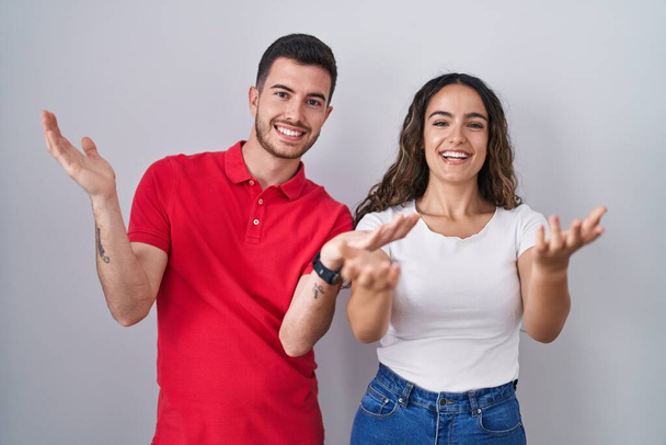 Junges hispanisches Paar, das vor isoliertem Hintergrund steht und fröhlich lächelnd Hände anbietet, die Hilfe und Akzeptanz geben.  - Foto, Bild