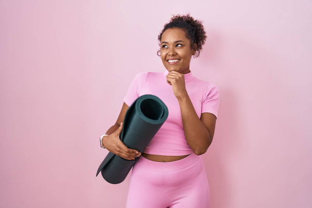 Junge hispanische Frau mit lockigem Haar, die eine Yogamatte über rosa Hintergrund hält, mit der Hand am Kinn über Fragen nachdenkend, nachdenklich. lächelndes und nachdenkliches Gesicht. Zweifelhaftes Konzept.  - Foto, Bild