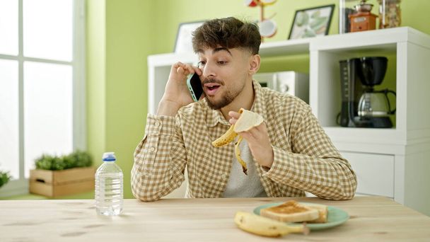 Young arab man having breakfast talking on smartphone at home - Фото, изображение