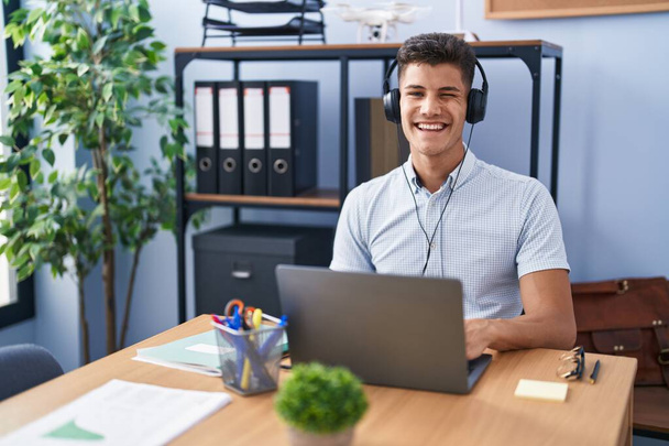 Νεαρός Ισπανός που εργάζεται στο γραφείο φορώντας ακουστικά να κλείνει το μάτι κοιτάζοντας την κάμερα με σέξι έκφραση, χαρούμενο και χαρούμενο πρόσωπο.  - Φωτογραφία, εικόνα