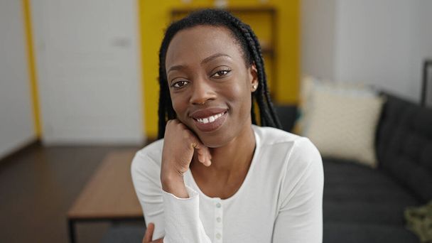 Африканская американка улыбается уверенно сидя дома на диване - Фото, изображение
