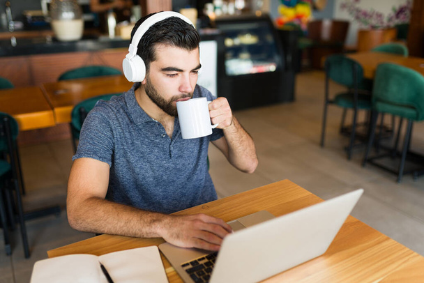 Jeune homme attrayant faisant du travail indépendant ou à distance sur l'ordinateur portable en utilisant le wifi tout en buvant du café au café  - Photo, image