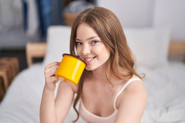 Молодая женщина пьет чашку кофе сидя на кровати в спальне - Фото, изображение