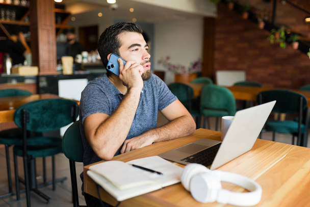 Зайнятий іспаномовний чоловік-підприємець у кав'ярні розмовляє під час робочого дзвінка по телефону і виконує віддалену роботу на ноутбуці - Фото, зображення
