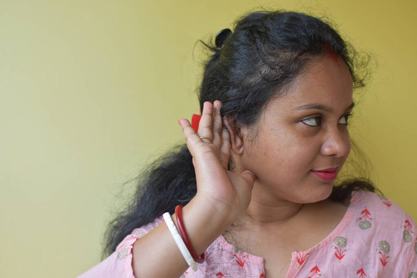 Любопытная индийская замужняя женщина держит за руку ухо и пытается услышать информацию, желтый фон. Селективный фокус на ухе. - Фото, изображение