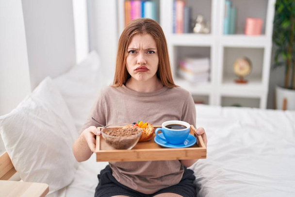 Mujer pelirroja con pijama sosteniendo la bandeja del desayuno deprimida y preocupada por la angustia, llorando enojada y asustada. expresión triste.  - Foto, imagen