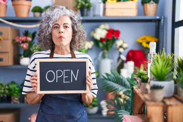 Mujer de mediana edad con pelo gris trabajando en floristería con signo abierto haciendo cara de pez con boca y ojos entrecerrados, loca y cómica.  - Foto, imagen
