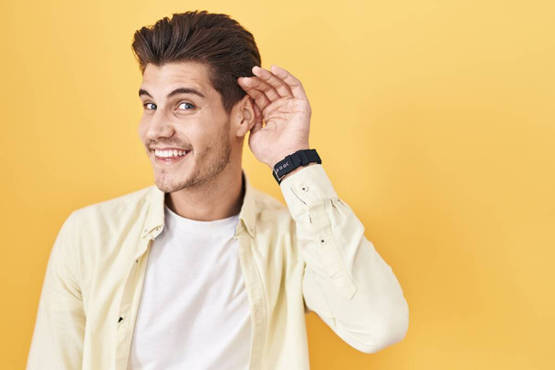 Молодий іспаномовний чоловік стоїть над жовтим тлом, посміхаючись рукою над вухом, слухаючи чутки або плітки. концепція глухоти
.  - Фото, зображення