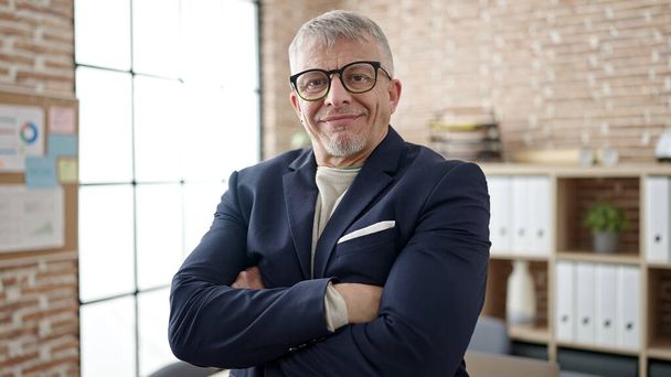 Hombre de pelo gris de mediana edad trabajador de negocios sonriendo confiado de pie con los brazos cruzados gesto en la oficina - Foto, imagen