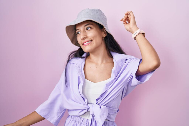Young hispanic woman standing over pink background wearing hat dancing happy and cheerful, smiling moving casual and confident listening to music  - Fotografie, Obrázek