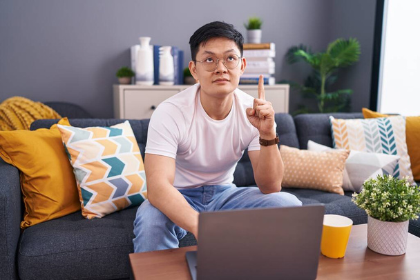 Junger asiatischer Mann mit Laptop sitzt zu Hause auf dem Sofa und zeigt traurig und aufgebracht nach oben, gibt mit den Fingern die Richtung vor, unglücklich und deprimiert.  - Foto, Bild