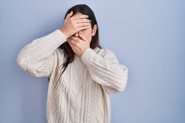 Young brunette woman standing over blue background covering eyes and mouth with hands, surprised and shocked. hiding emotion  - Fotografie, Obrázek