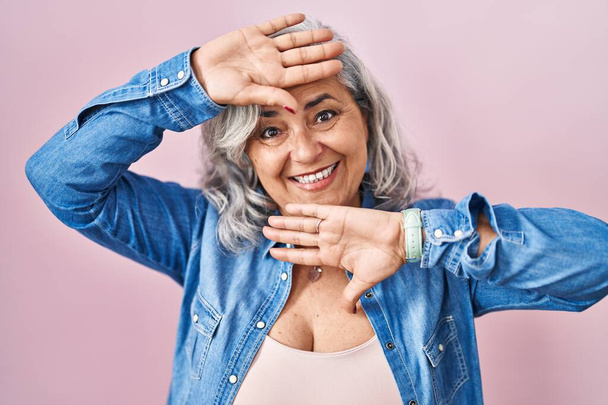 Жінка середнього віку з сірим волоссям, що стоїть над рожевим тлом, усміхнений веселий граючи зазирнути в бу з руками, що показують обличчя. здивований і вийшов
  - Фото, зображення