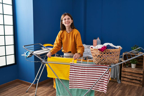 Junge blonde Frau lächelt selbstbewusst, hängt Kleidung an Wäscheleine in der Waschküche - Foto, Bild