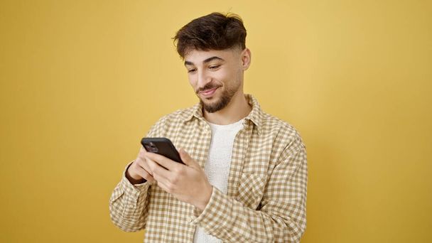 Молодой араб уверенно улыбается, используя смартфон на изолированном желтом фоне - Фото, изображение