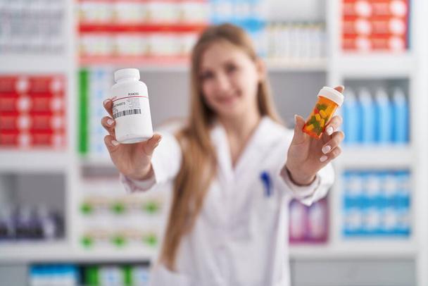 Giovane farmacista donna caucasica sorridente fiducioso tenendo bottiglie pillole in farmacia - Foto, immagini
