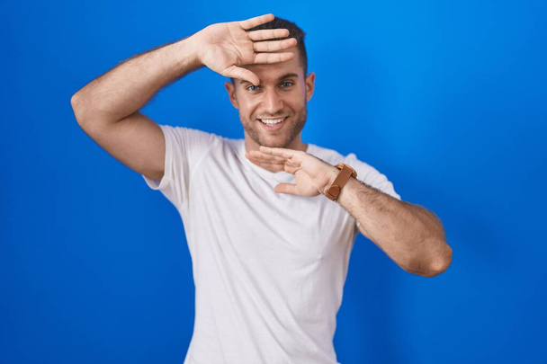 Young caucasian man standing over blue background smiling cheerful playing peek a boo with hands showing face. surprised and exited  - Photo, image