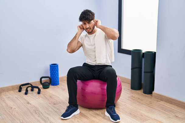 Spaanse man met baard zittend op pilatesballen in de yogakamer die oren bedekt met vingers met geïrriteerde uitdrukking voor het lawaai van luide muziek. doof concept.  - Foto, afbeelding