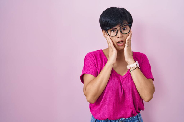 Young asian woman with short hair standing over pink background afraid and shocked, surprise and amazed expression with hands on face  - Foto, afbeelding