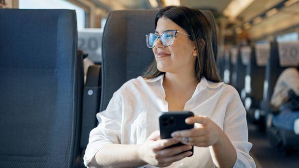 列車のワゴンの中に座っているスマートフォンを使用して微笑む若い美しいヒスパニックの女性 - 写真・画像